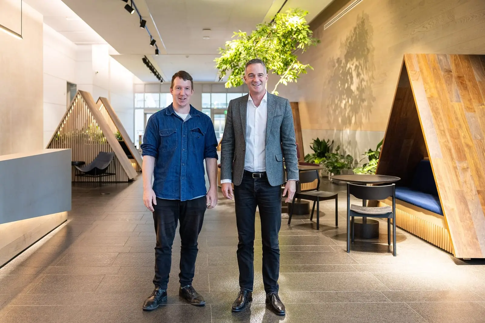 Two men stand side by side in a bright office lobby with wooden booths and a large indoor tree.