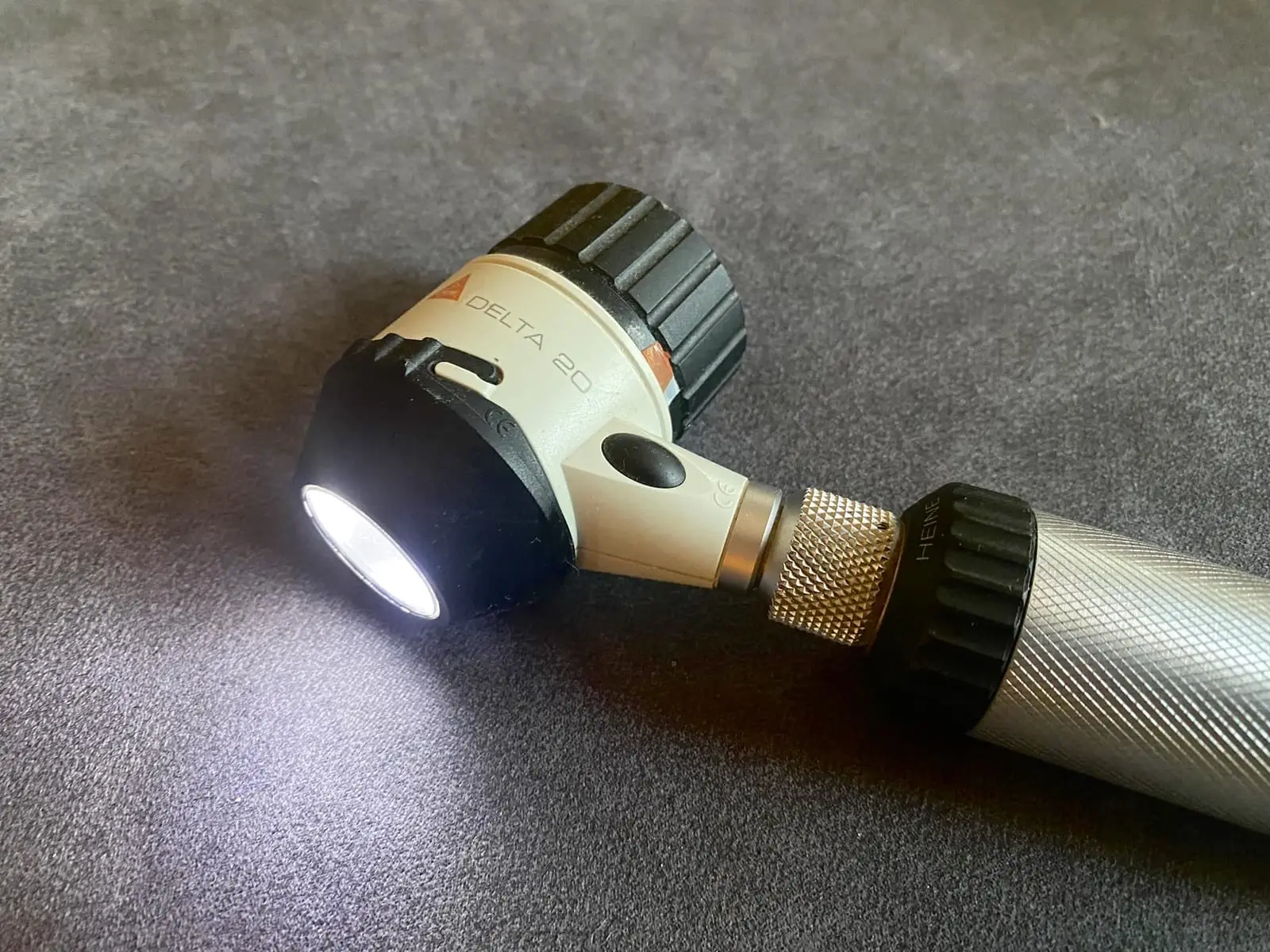A handheld dermatoscope with its light switched on, lying on a grey surface.