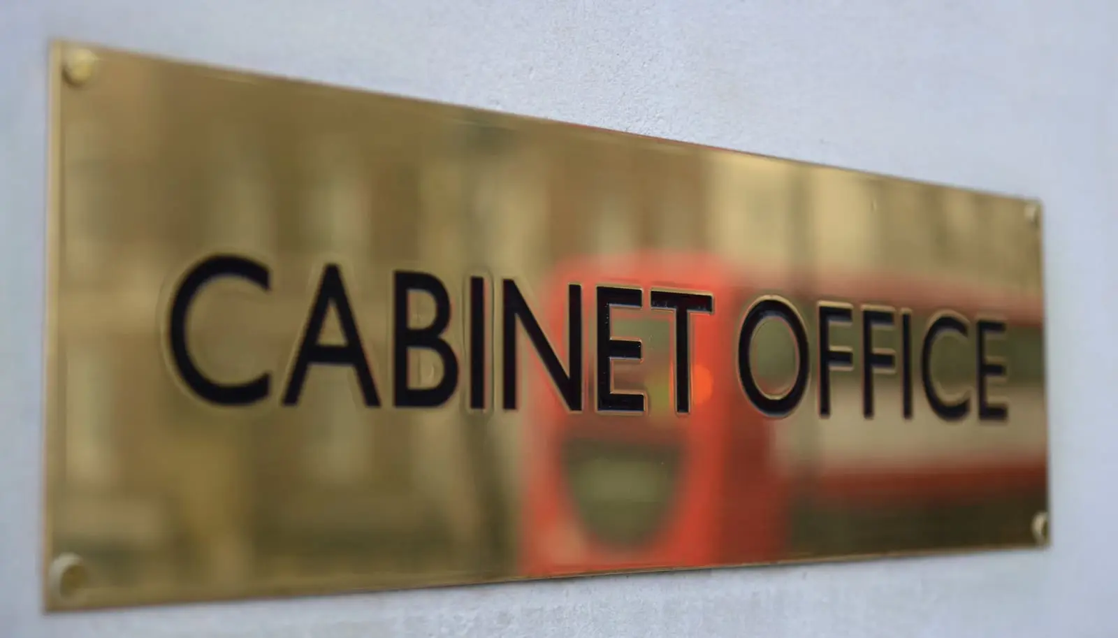 A brass Cabinet Office sign on a stone wall in Whitehall, London, with a red bus reflected in it.